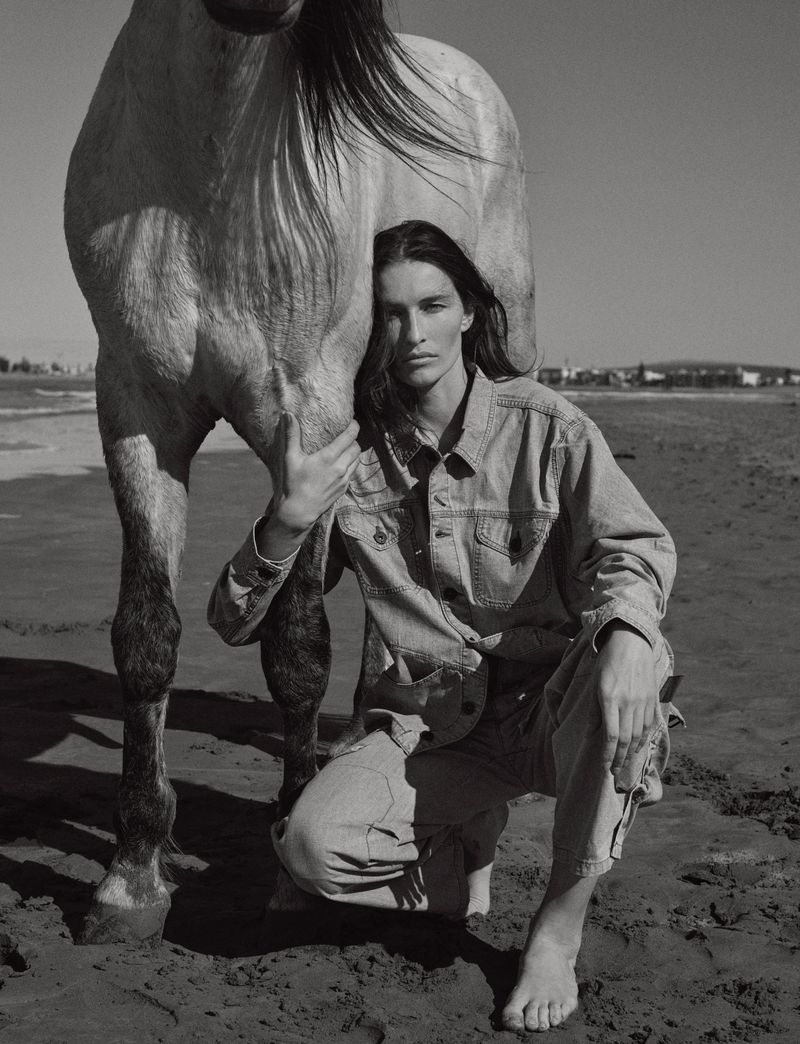 A woman in a denim outfit kneels barefoot on a sandy beach, tenderly embracing a horse in a black-and-white editorial photograph.