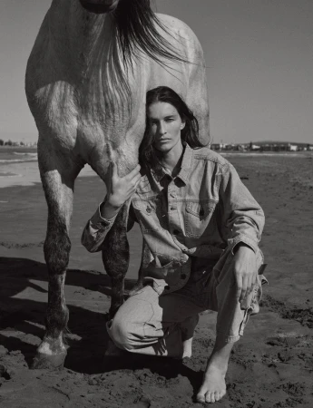 A woman in a denim outfit kneels barefoot on a sandy beach, tenderly embracing a horse in a black-and-white editorial photograph.