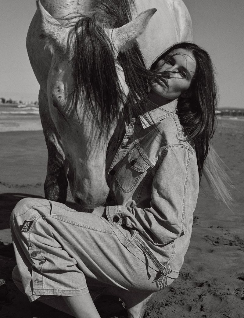 Monochrome portrait of a woman crouching with a white horse on a beach with an ocean background under a clear sky.