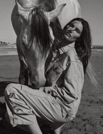 Monochrome portrait of a woman crouching with a white horse on a beach with an ocean background under a clear sky.