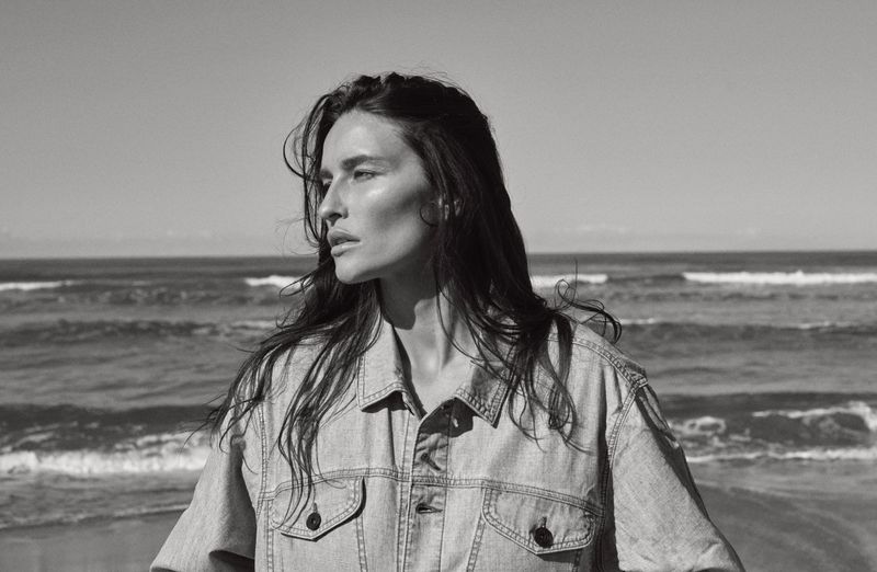 Black and white portrait of a woman with long hair, wearing a denim jacket, standing on a beach with ocean waves in the background.