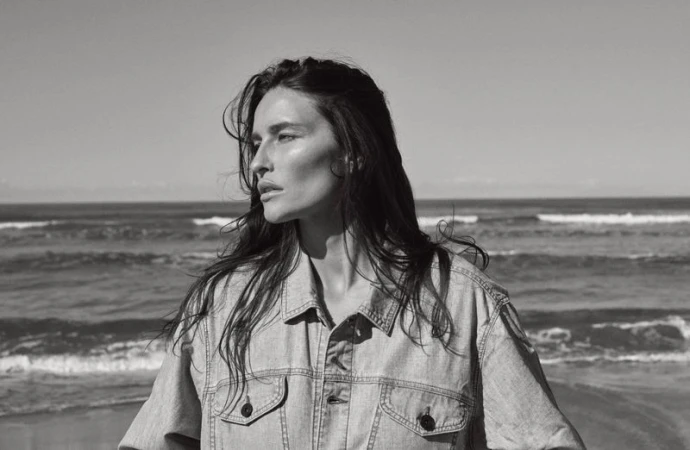 Black and white portrait of a woman with long hair, wearing a denim jacket, standing on a beach with ocean waves in the background.