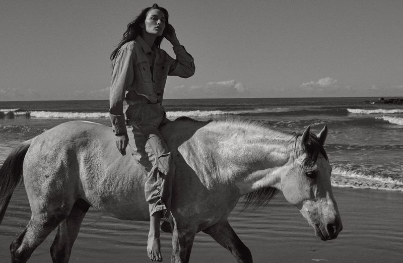 Monochrome fashion editorial image of a model riding a white horse along a sandy beach near the ocean.