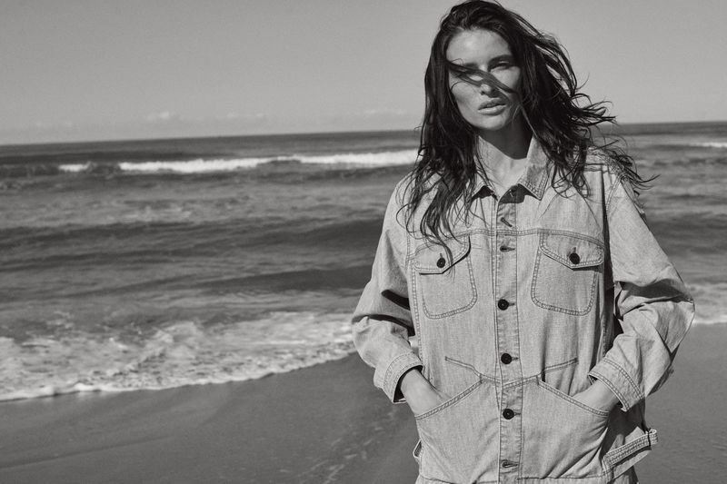 Adult female in a light denim jacket poses on a beach in a black-and-white outdoor shot with the ocean and sky as a backdrop.
