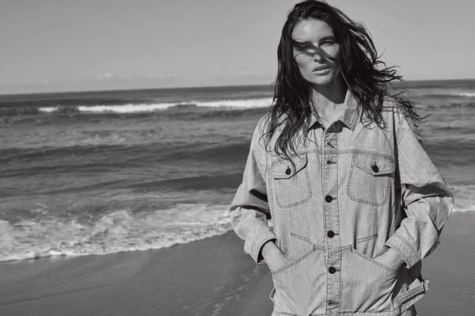 Adult female in a light denim jacket poses on a beach in a black-and-white outdoor shot with the ocean and sky as a backdrop.