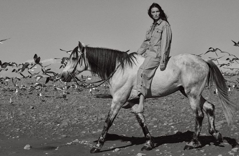 Black and white image of a woman riding a horse on a beach with a flock of seagulls flying and standing in the background.