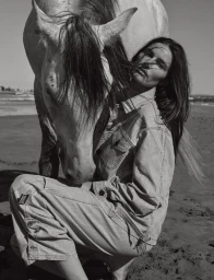 Monochrome fashion portrait of a model in denim crouching next to a horse on a sandy beach
