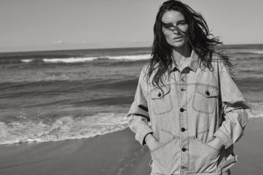 Black and white portrait of a woman wearing a denim jacket standing by the ocean on a sandy beach.