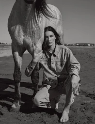 Model in denim outfit crouching next to a white horse on a sandy beach