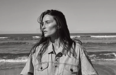 Black and white portrait of a woman wearing a denim jacket standing against a coastal ocean backdrop.