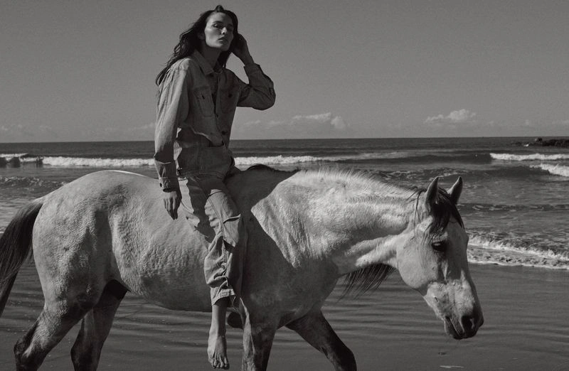 Monochrome fashion editorial image of a model riding a white horse along a sandy beach near the ocean.
