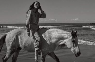 Monochrome fashion editorial image of a model riding a white horse along a sandy beach near the ocean.