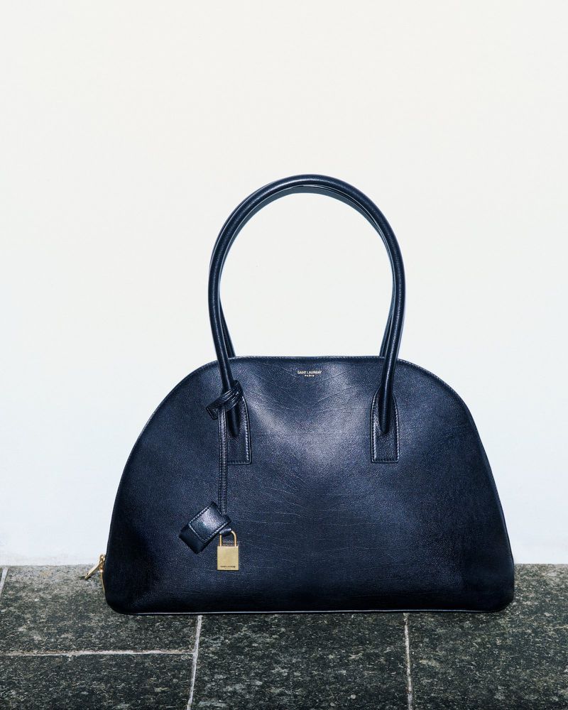 Black leather Saint Laurent Dome handbag with top handles, padlock, and gold zipper, placed on a tile surface against a white background.