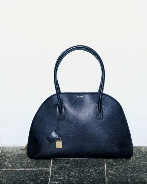 Black leather Saint Laurent Dome handbag with top handles, padlock, and gold zipper, placed on a tile surface against a white background.