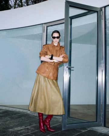 A woman poses in a caramel leather jacket, tan skirt, and red boots against a modern building with glass doors, capturing a fashion-forward look.