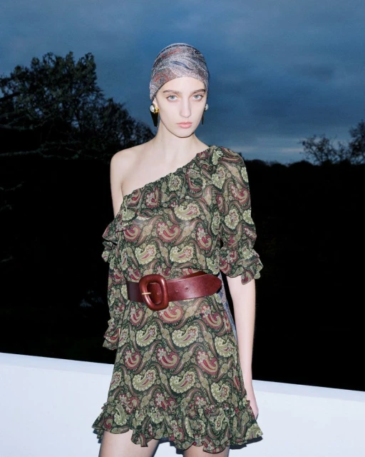 A woman poses outdoors in a one-shoulder paisley dress with a wide belt and matching headscarf against a dark background with trees.