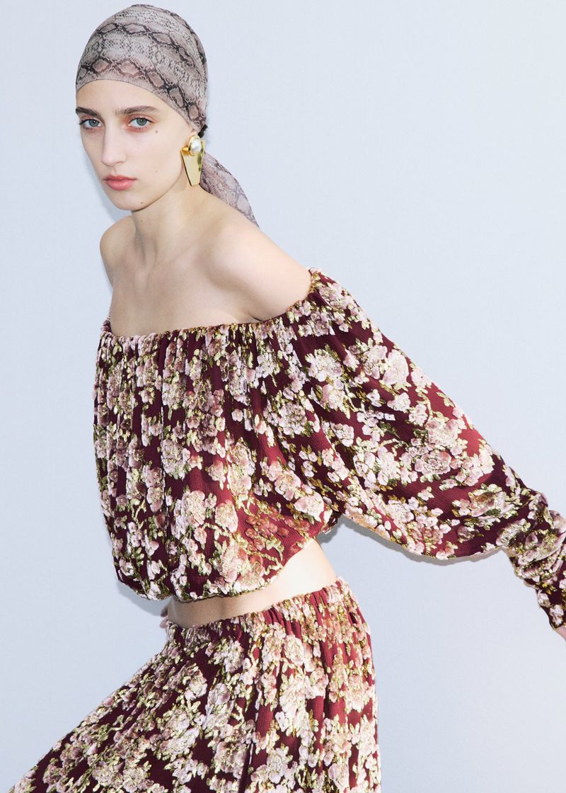 Model wearing a burgundy floral off-the-shoulder top and matching skirt with a patterned headscarf against a white background.