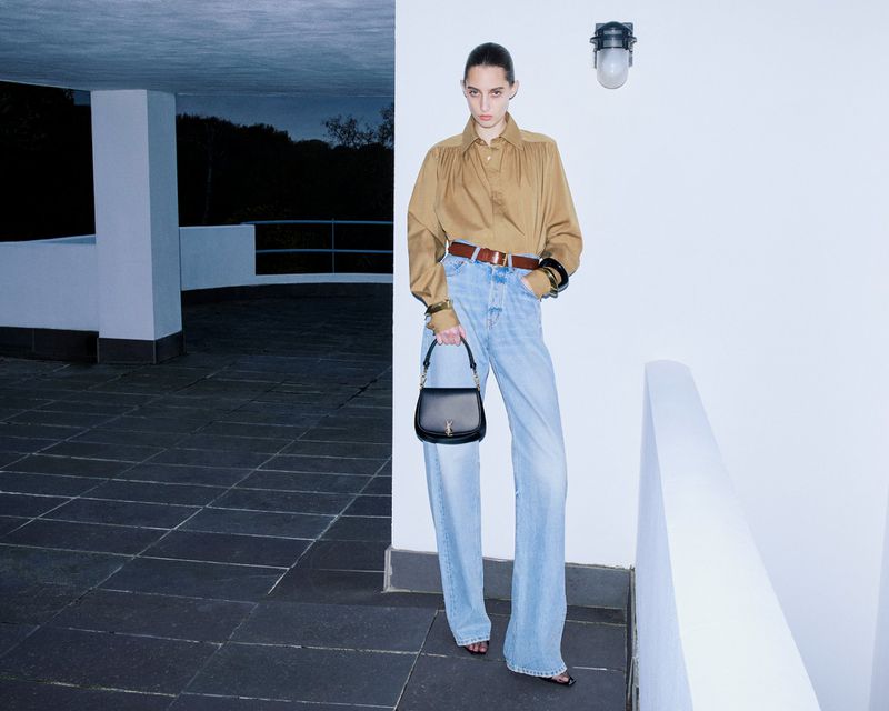 Full-length shot of a woman in a tan blouse and light-wash jeans holding a black purse against a white architectural backdrop.