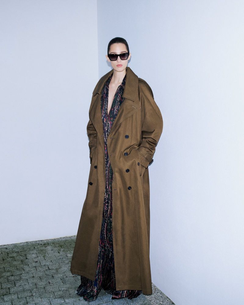 Model wearing dark sunglasses and a long brown trench coat standing against a pale corner wall