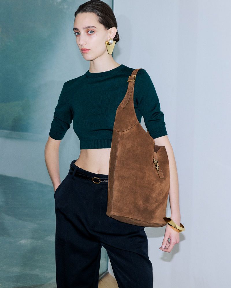 Fashion shot of a woman in a green top and dark pants with a brown suede purse and gold jewelry posing in front of a wall.