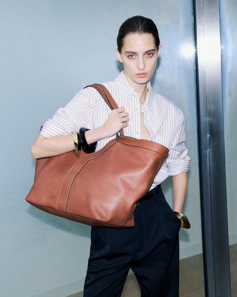 Model wearing a striped shirt and dark trousers holding a large brown leather tote bag against a neutral wall