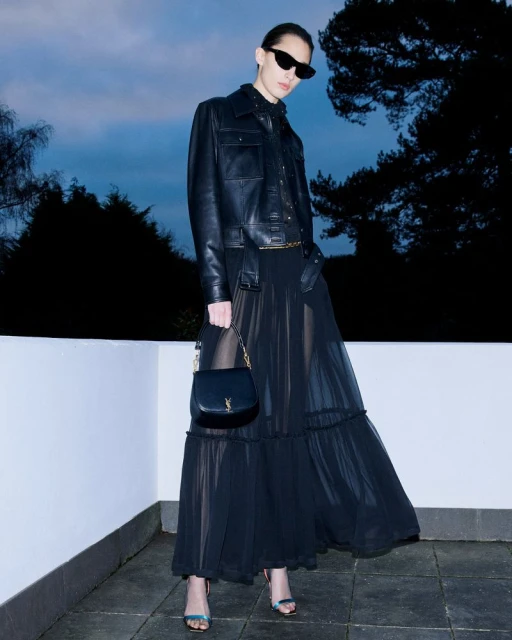 Elegant woman in a black leather jacket and flowing black dress, accessorized with sunglasses and a handbag, standing outdoors at dusk.