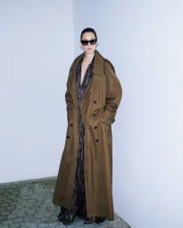 Model wearing dark sunglasses and a long brown trench coat standing against a pale corner wall