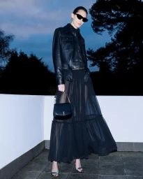 Model in black sunglasses wearing a leather jacket and sheer long skirt holding a handbag on a rooftop at dusk