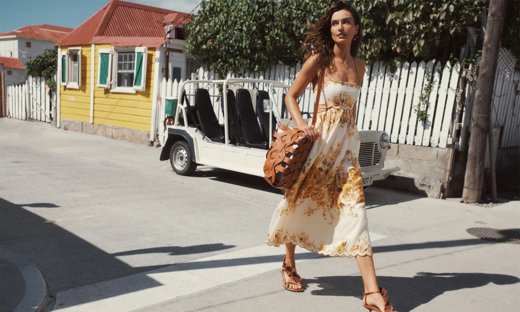 A woman in a floral dress crossing a street in a sunny, tropical setting with a small electric vehicle and colourful buildings.
