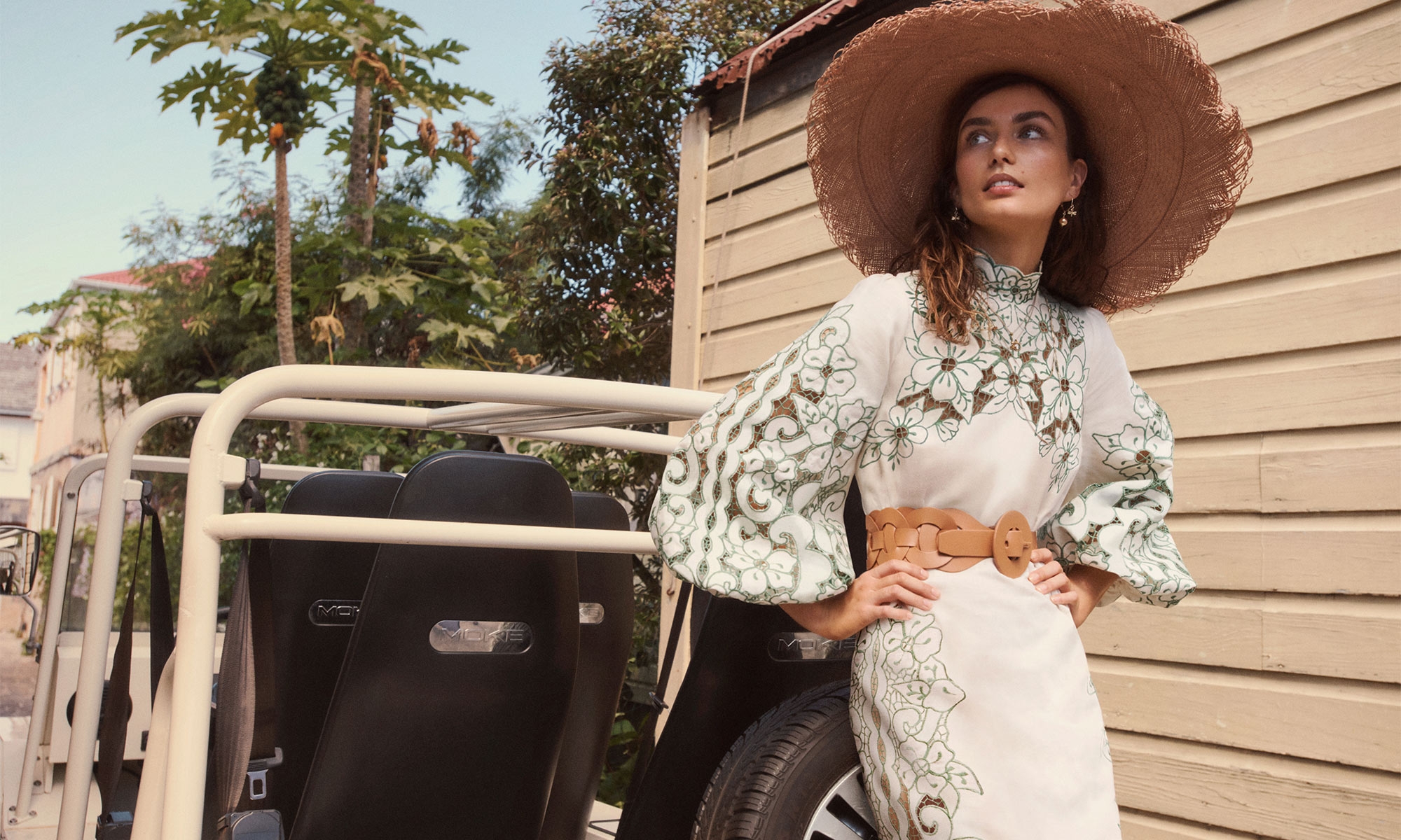 Model wearing a white patterned dress and wide-brim straw hat leaning against a vehicle exterior