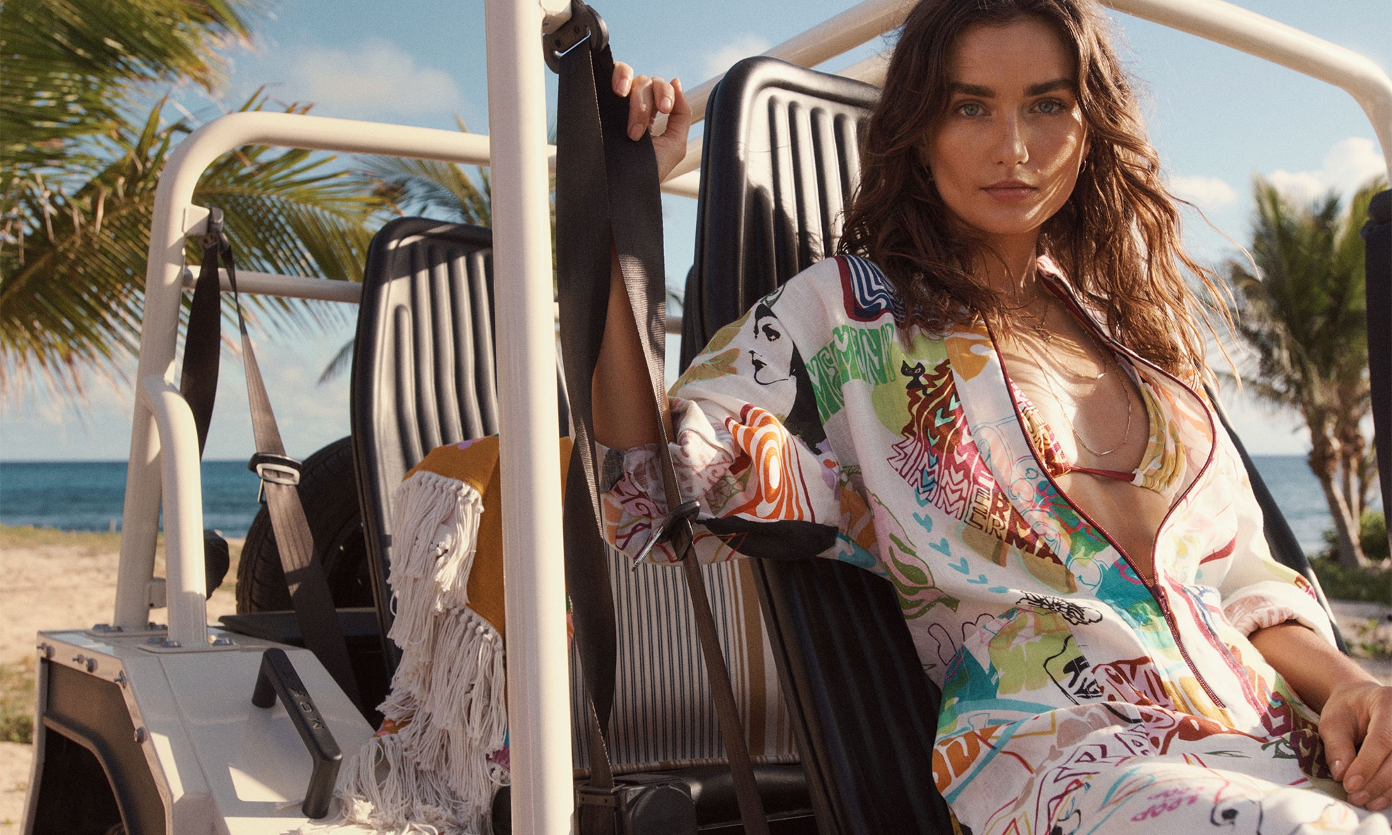 Adult female in colorful patterned shirt sits inside a white vehicle with a fringe bag, ocean, and palm trees in the background.