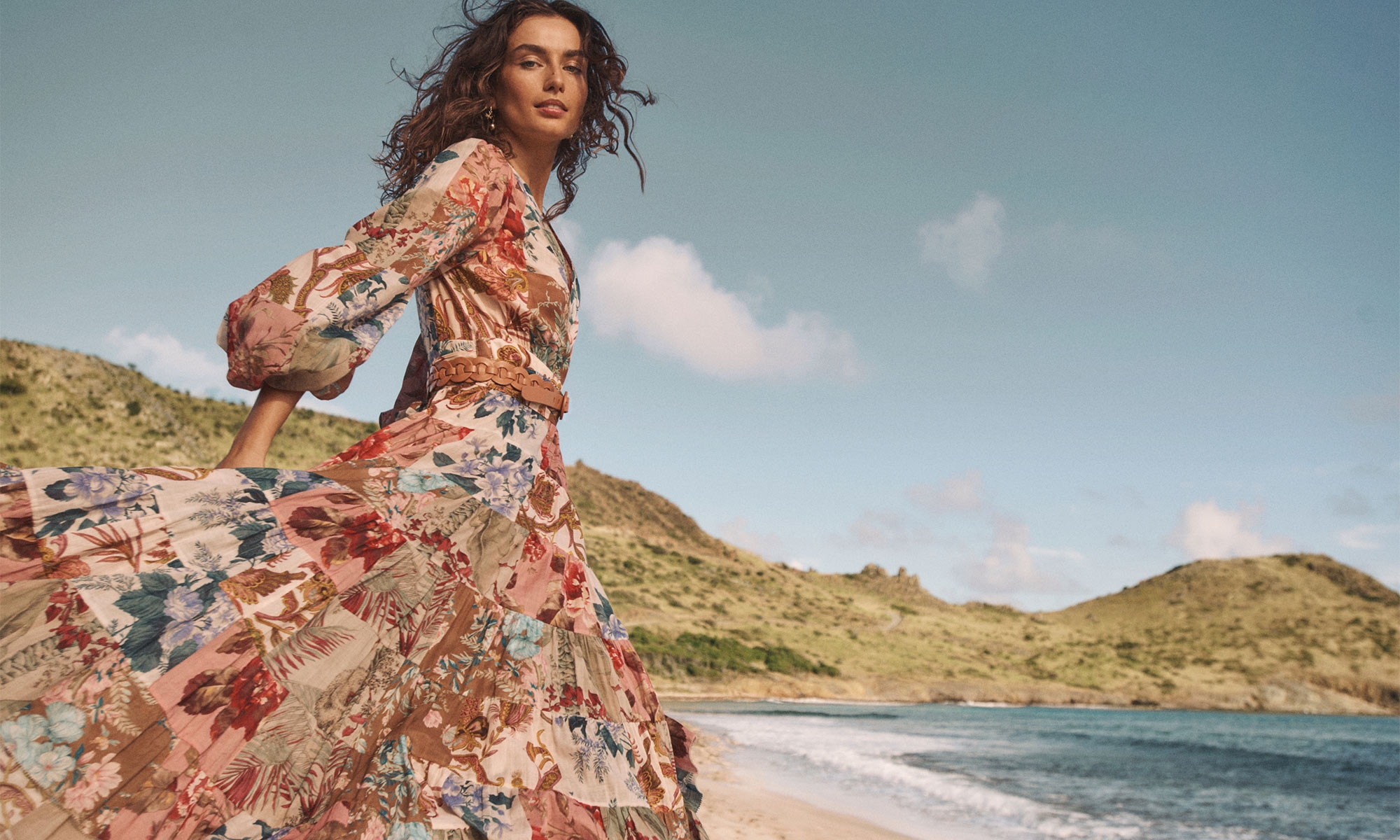 Model in a floral print maxi dress on a sandy beach with rolling coastal hills and ocean in the background