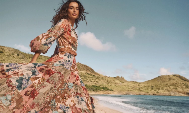 Model in a floral print maxi dress on a sandy beach with rolling coastal hills and ocean in the background