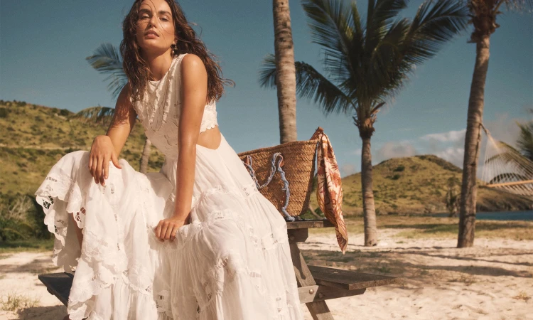 Woman in a white lace dress seated on a wooden bench outdoors near palm trees and a sandy beach
