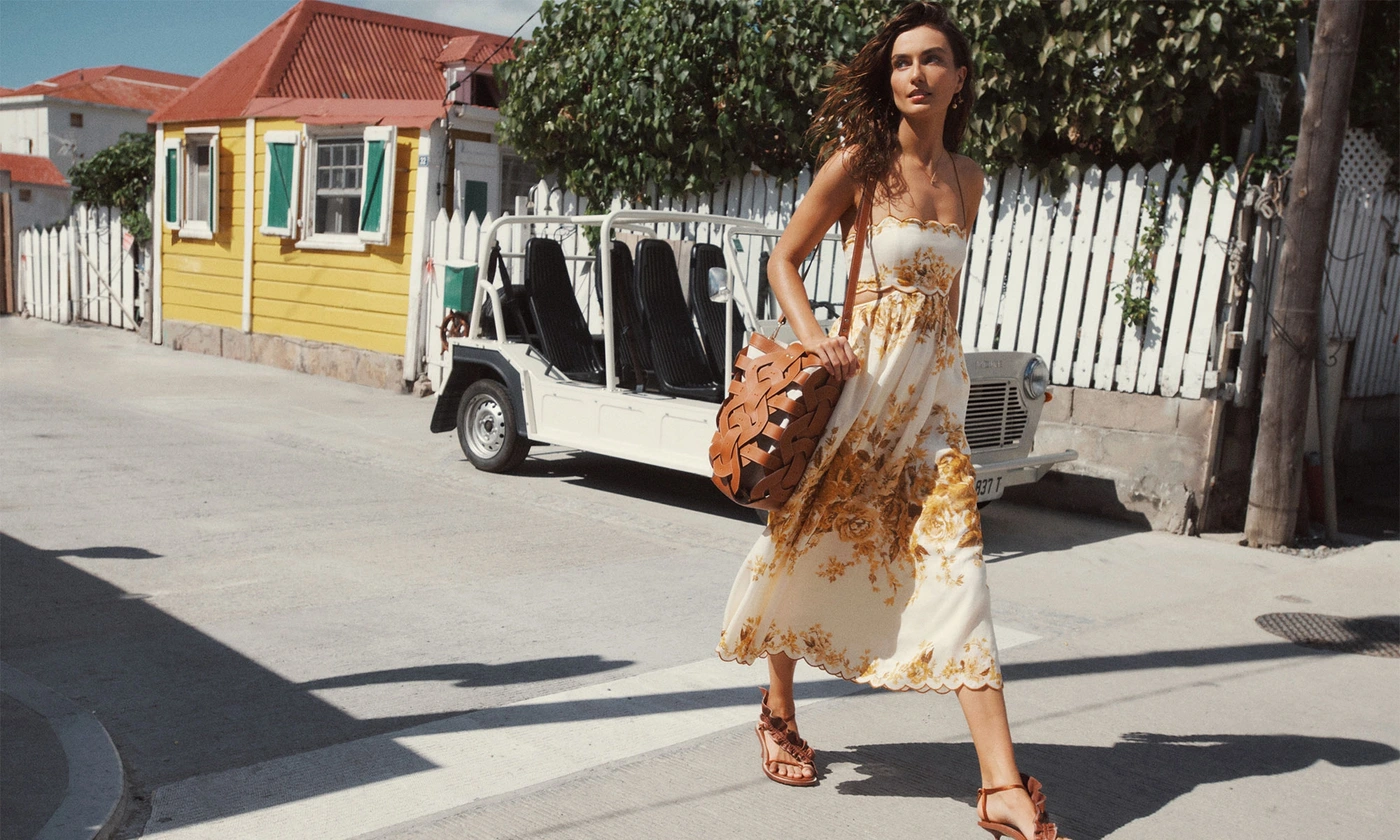 Woman walking on a sunny street wearing a printed summer dress and carrying a woven leather bag, with a yellow building behind.