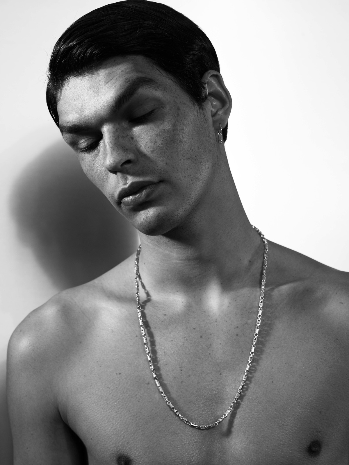 Monochrome portrait of a young adult male with his eyes closed, wearing a chain necklace against a plain white background.