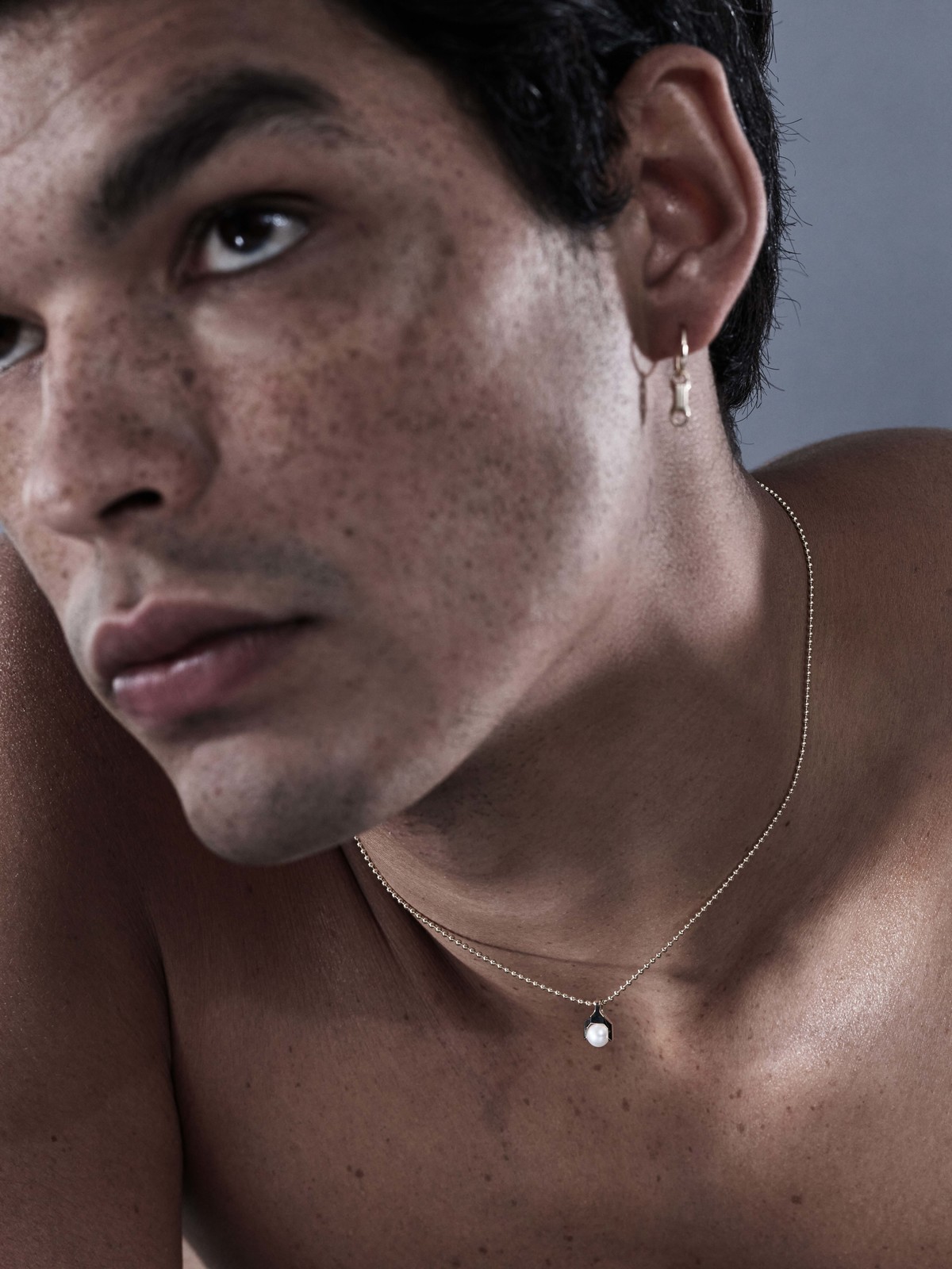Close-up portrait of a man wearing delicate gold jewelry including earrings and a pendant necklace for Sarah & Sebastian.