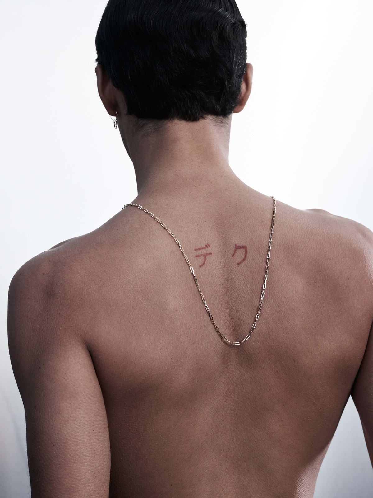 Back view of an adult with fair skin and short dark hair wearing a gold necklace and earring, set against a white backdrop.