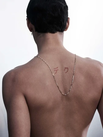 Back view of an adult with fair skin and short dark hair wearing a gold necklace and earring, set against a white backdrop.