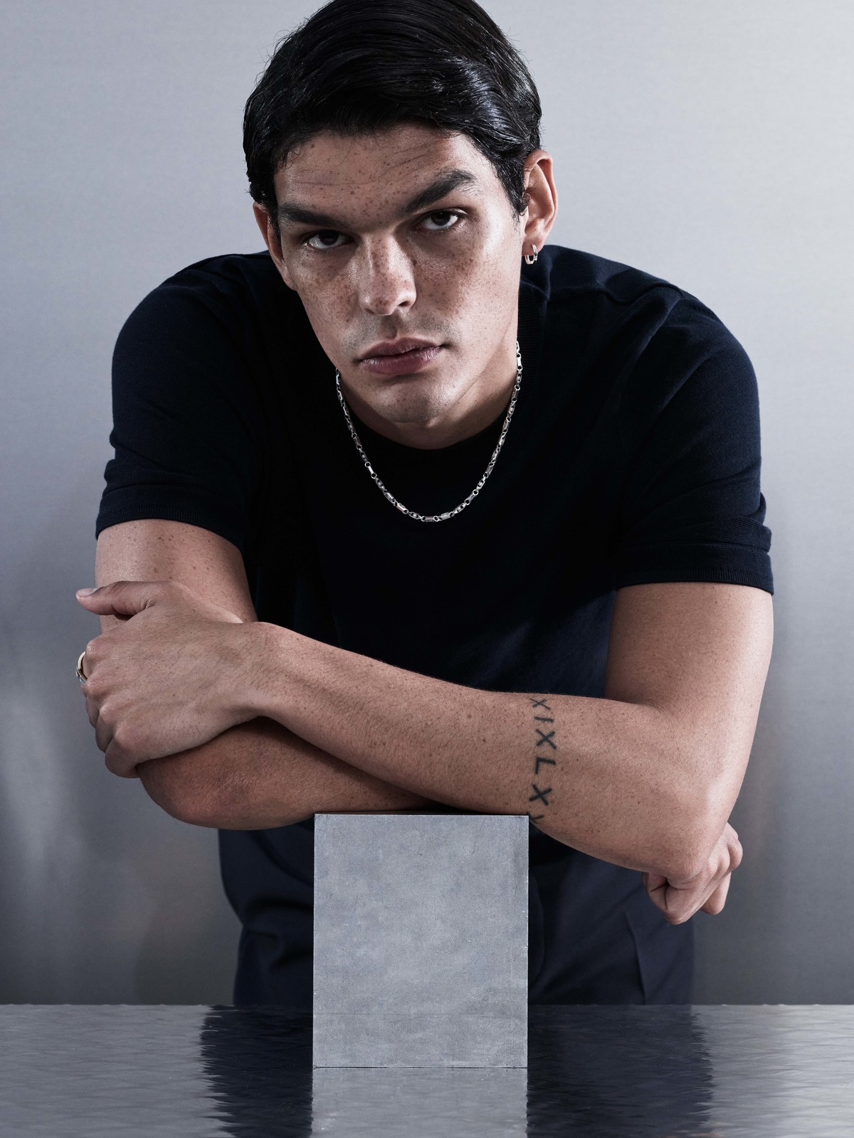 Man in a black shirt poses in a studio with arms crossed on a reflective surface, wearing a silver chain and sporting a tattoo.