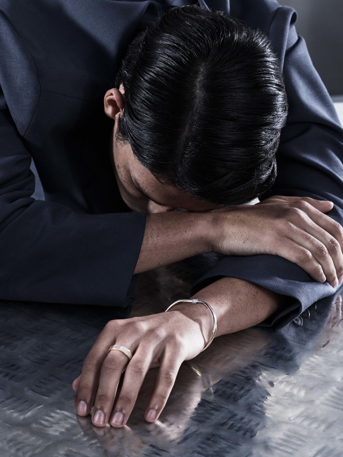 Man with head down on metallic surface, wearing a navy suit and gold and silver bracelets, indoor setting.
