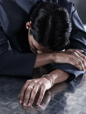 Man with head down on metallic surface, wearing a navy suit and gold and silver bracelets, indoor setting.