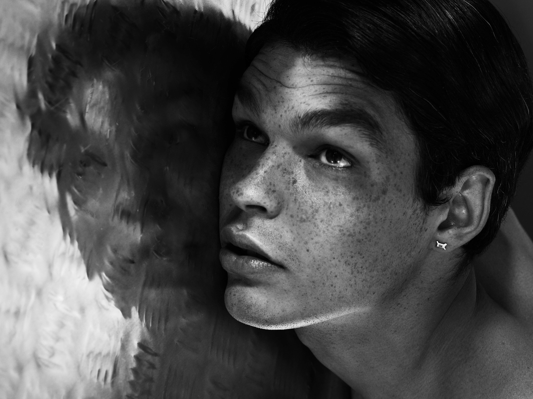 Close-up portrait of a freckled young adult man with an earring, near textured feathers, black and white, indoor, looking up.