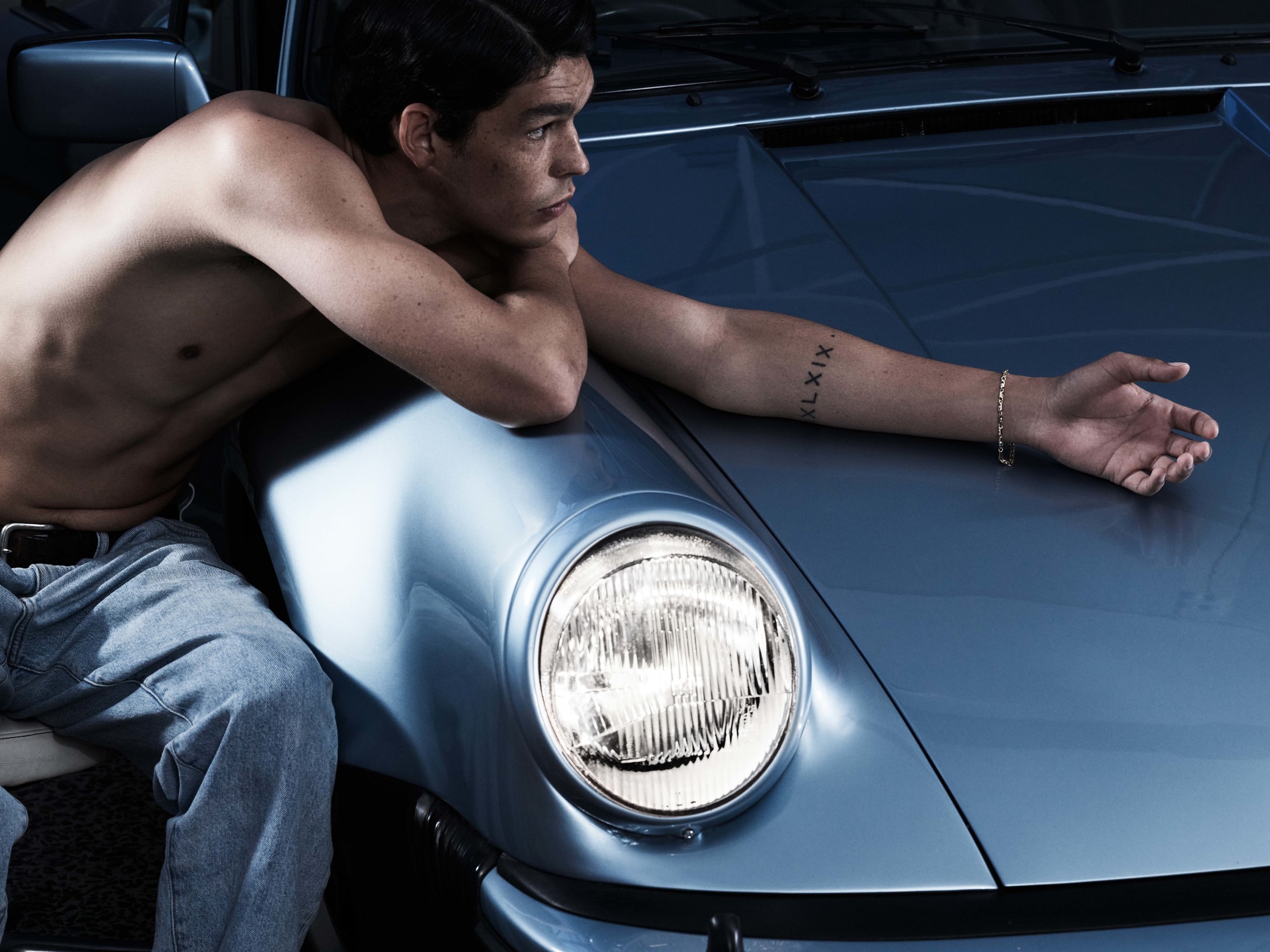 A man leans against a blue sports car, showcasing a casual yet stylish aesthetic with a focus on automotive elegance and relaxed fashion.