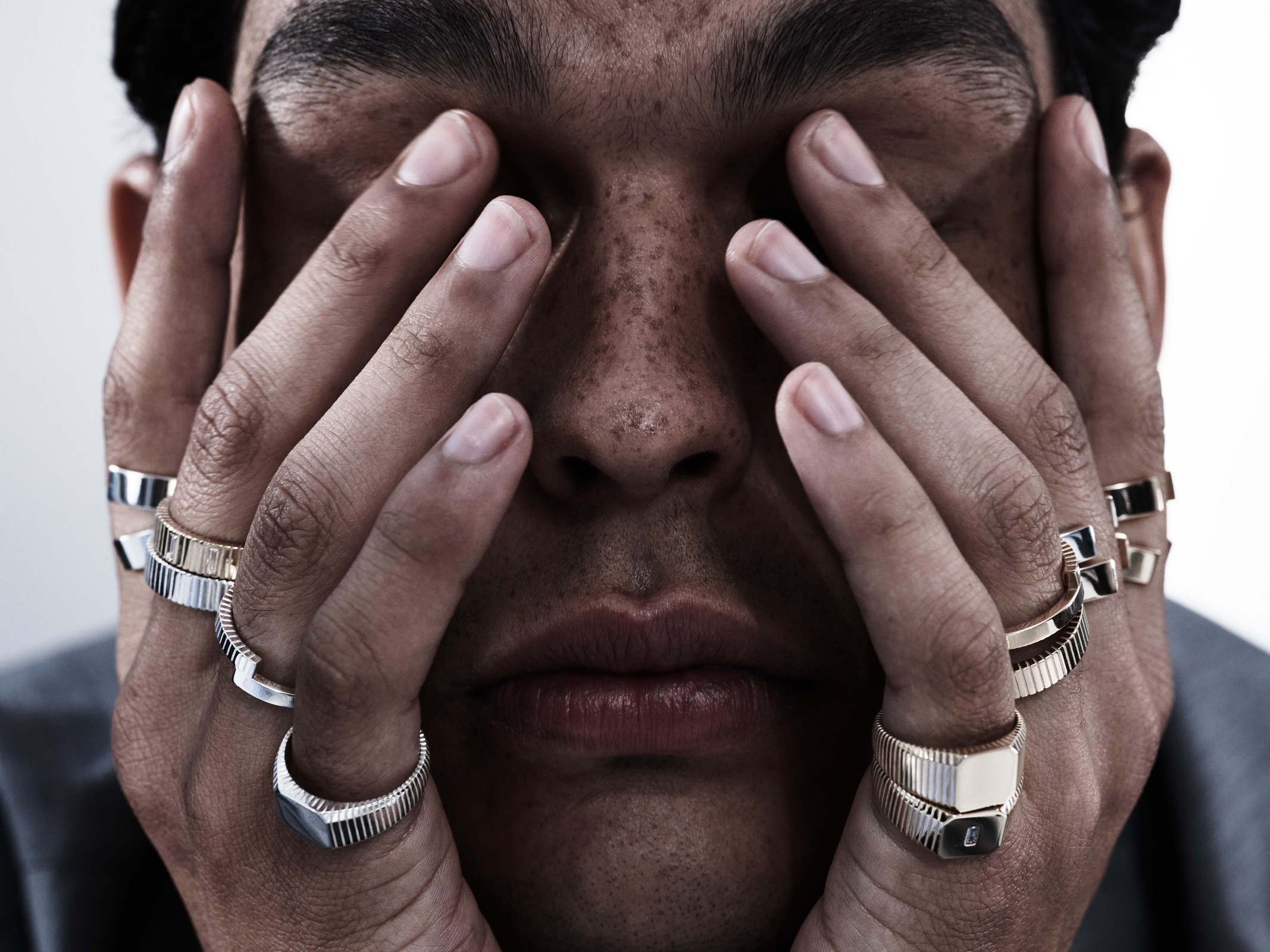 Close-up shot of a person with multiple rings, covering their face with their hands in an indoor setting against a neutral background.