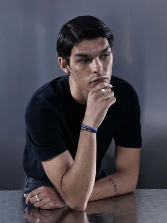 Close-up portrait of a man leaning on a reflective surface wearing a black t-shirt, rings, and bracelets for Sarah & Sebastian.