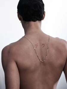 Rear view of a topless man wearing a gold chain necklace and a small earring against a plain white studio background.