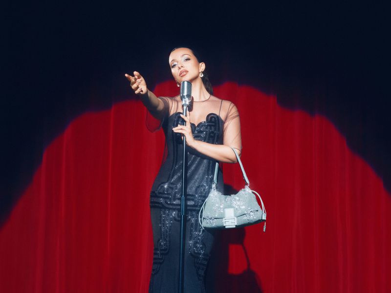 Elegant woman in black dress performs on stage, holding a vintage microphone, with a red curtain backdrop and a blue beaded purse.