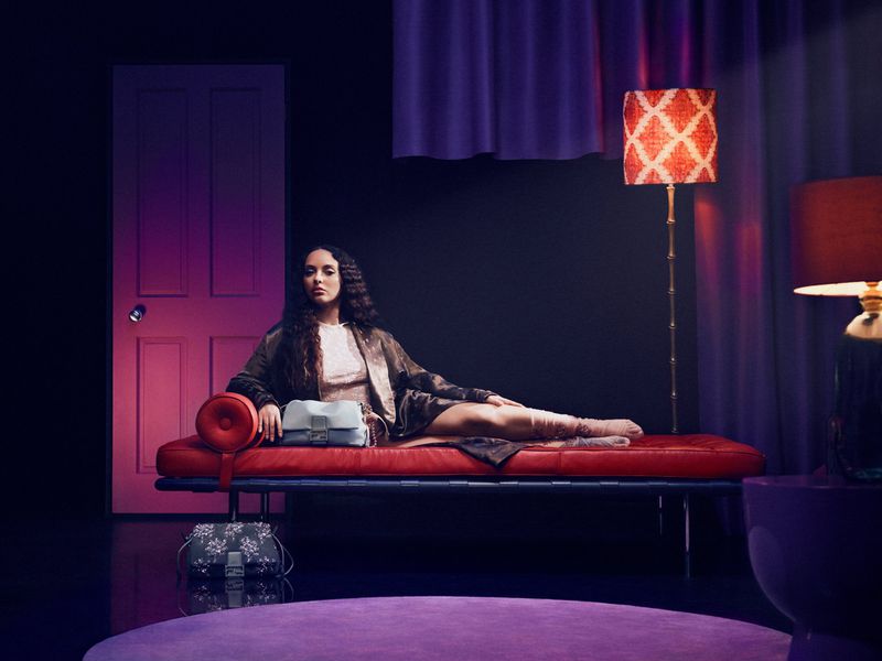 A woman lounges on a red chaise in a studio setting, holding a light blue handbag, with a dark background and ambient lighting.