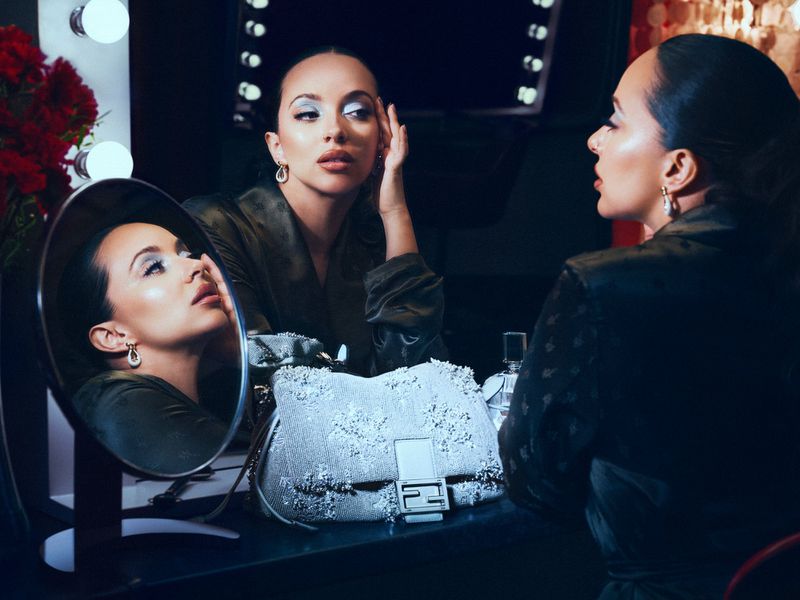 Woman looking in mirror with designer handbag on vanity. She wears a dress and earrings in a dark setting with soft lighting.